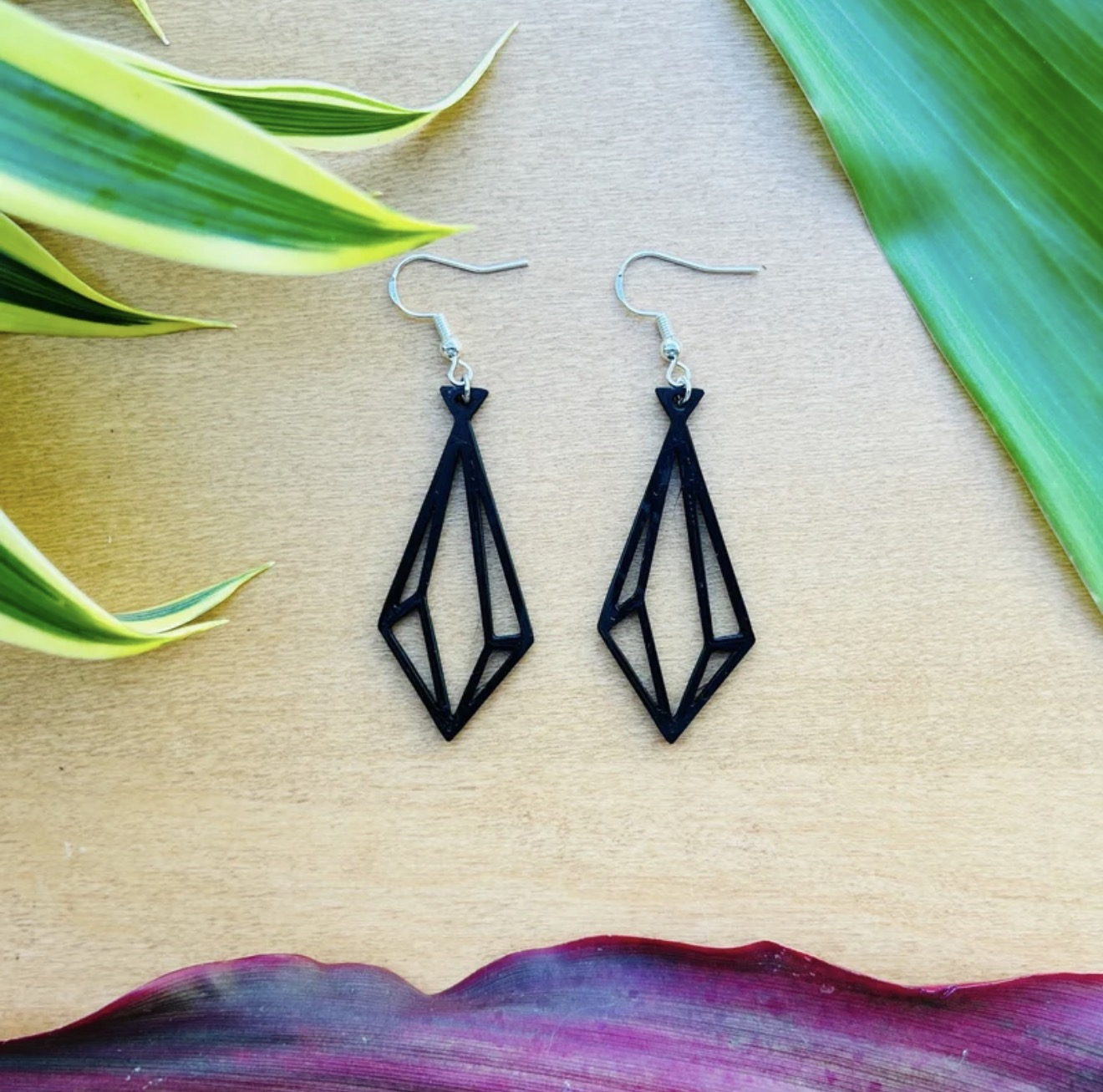 Black 3D printed diamond-shaped earrings displayed on a wood surface with leaves.